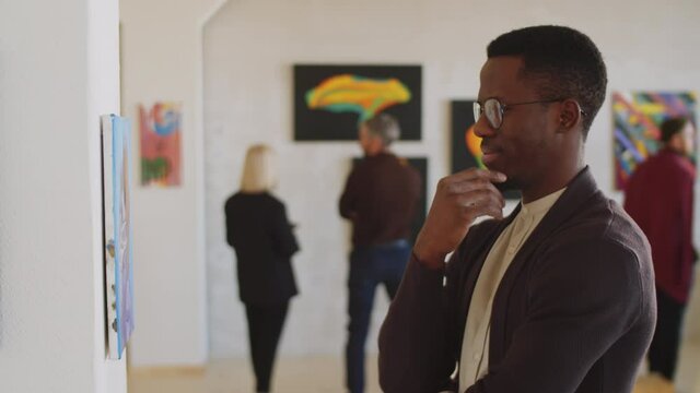 Portrait of young Afro-American man examining painting on the wall and then posing for camera and smiling while standing in art gallery