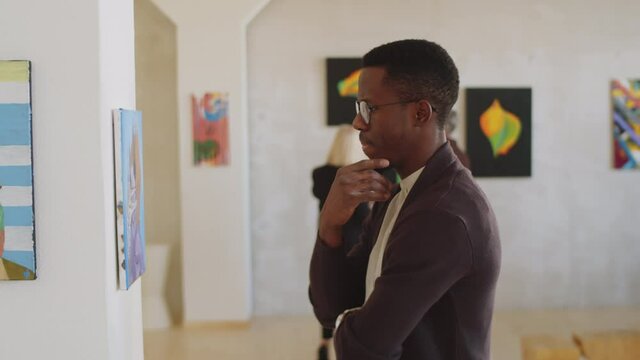 Side view of young Afro-American man rubbing his chin and looking at painting on the wall pensively while visiting modern art exhibition in gallerySide view of young Afro-American man rubbing his chin