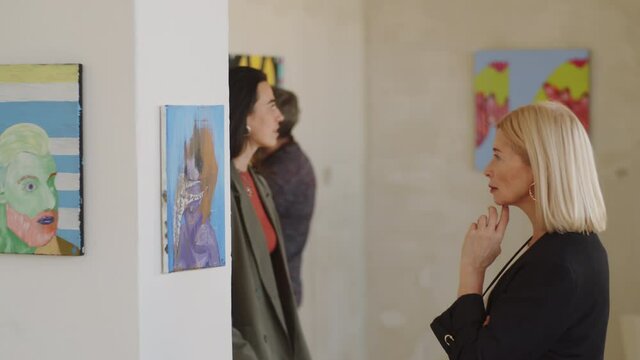 Side view of beautiful middle aged woman standing with hand under chin in art gallery and examining abstract painting on the wall