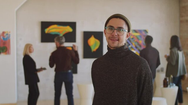 Portrait of young Caucasian man watching painting on the wall and then looking at camera and smiling while visiting art gallery