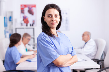 Smiling nurse in wearing blue uniform in hospital conference room looking at camera with medical staff in the background. Friendly medical practitioner in clinic meeting room, robe, specialist.