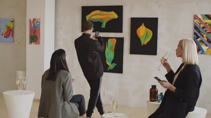 Two female gallery managers having champagne and using smartphones in exhibition hall while visitors examining paintings