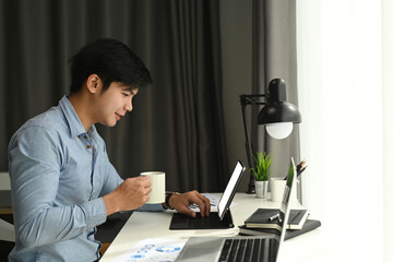 Side view of a graphic designer is working with computer tablet and drinking coffee at his workspace in the morning.