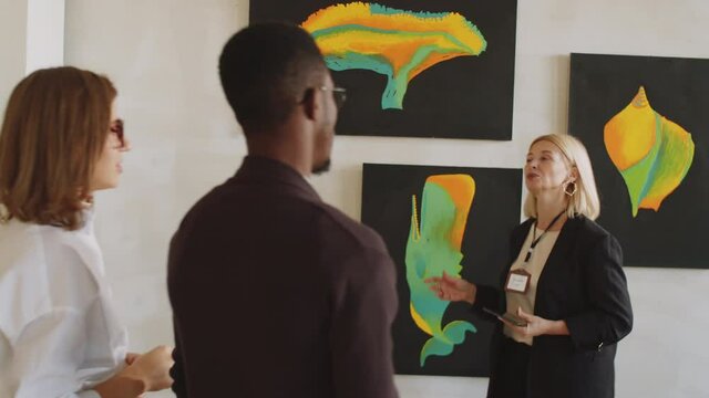 Elegant middle aged female guide holding digital tablet and telling about artworks on walls to visitors while showing them around exhibition in gallery