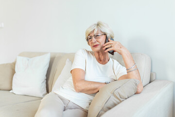 elderly woman grandmother with gray hair sits at home on the couch using mobile phone, a telephone conversation to hear the bad news. Emotion fear scare.