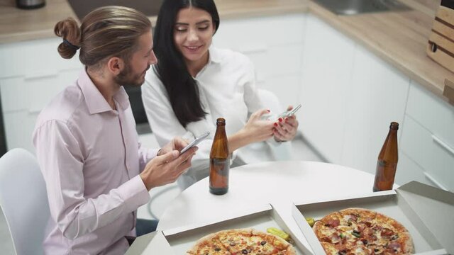 Home party, a young couple sitting at table in kitchen and taking pictures of pizza on smartphones, taking food photo with a mobile phone camera.