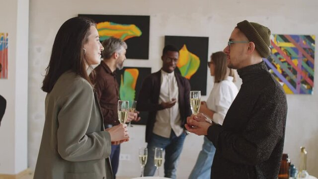 Young man and woman holding flutes with champagne and discussing something at exhibition opening party in art gallery