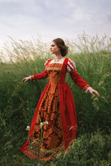 Obraz na płótnie Canvas beautiful young woman in a long red medieval dress is standing in the grass in the field under the cloudy sky, fantasy princess