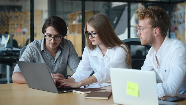 Modern young business people in glasses sitting by the table and working and using laptop together.