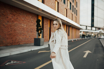 Fototapeta premium Serious trendy young girl in a beige trench coat in an elegant jeans in trench coat posing outdoors on a road. 