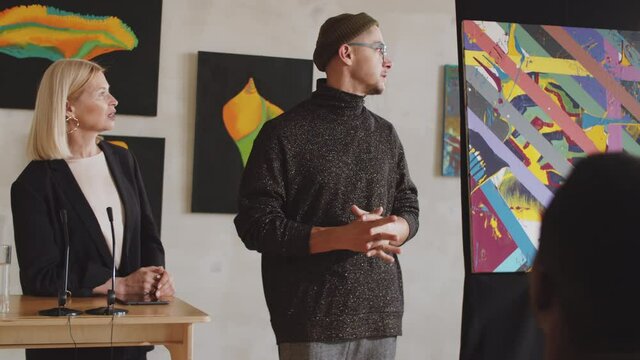 Young trendy male artist speaking with female auctioneer and giving presentation about his abstract painting to audience during art auction in gallery