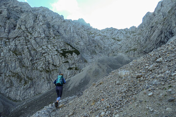 Obraz premium A woman with backpack hiking through steep and high mountain crater in Austrian Alp. The sun shines behind the mountain wall. The whole area is covered with shadow. Steep and narrow path along.