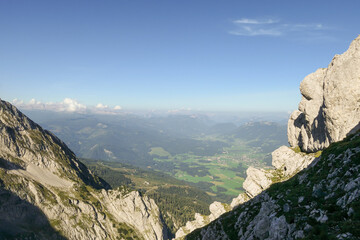 A panoramic view from an Alpine top on a vast valley. There are sharp mountains and high peaks around. The Alpine slopes are almost barren. Lush green valley. Bright day. Serenity and freedom.