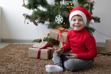 Caucasian boy wearing a red santa claus hat. sitting near the Christmas tree a box with a red bow next to it.