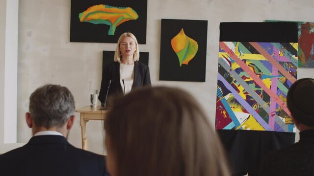 Beautiful mid-aged female auctioneer standing at rostrum, holding digital tablet and presenting abstract artwork to audience during auction