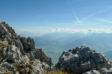 A panoramic view from an Alpine top on a vast valley. There are sharp mountains and high peaks around. The Alpine slopes are almost barren. Lush green valley. Bright day. Serenity and freedom.