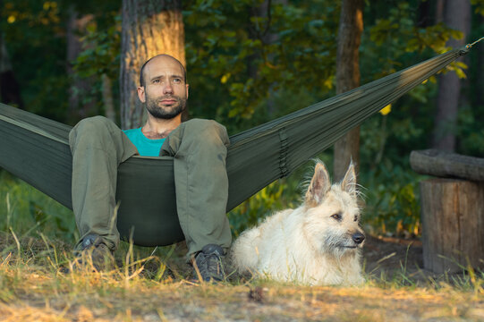Closeup portrait camping and resting man with white dog lying in hammock in green forest