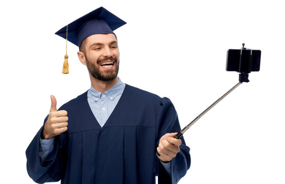 education, graduation and people concept - happy smiling male graduate student in mortar board and bachelor gown taking picture with smartphone on selfie stick over white background