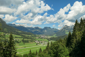 Panoramic view on a vast Alpine valley. There are sharp mountains and high peaks around. Small town at the bottom. The Alpine slopes are almost barren. Lush green valley. Bright day. Idyllic landscape