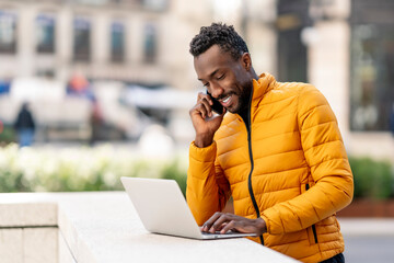 Happy african man using laptop and talking on smartphone on balcony in city
