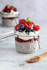 Two mason jars with chia pudding and fresh berry fruits and mint on a white background with a wooden spoon with chia seeds in front