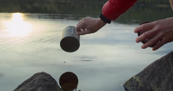 Woman in puffy jacket at campground scoop water with cup from sweet drinking clean lake. Camping lifestyle, cook on open fire. Boiling water on fireplace. Drink clean norwegian mountain glacier water