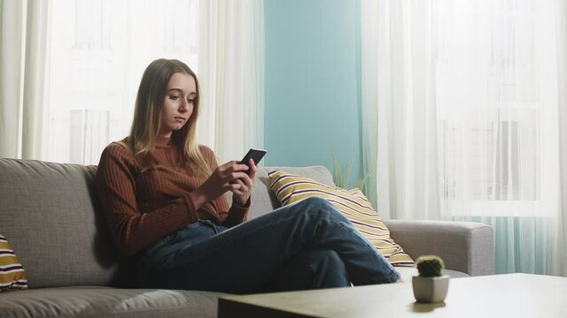 Young lady in blue jeans and brown sweater sitting on sofa and picks up phone.