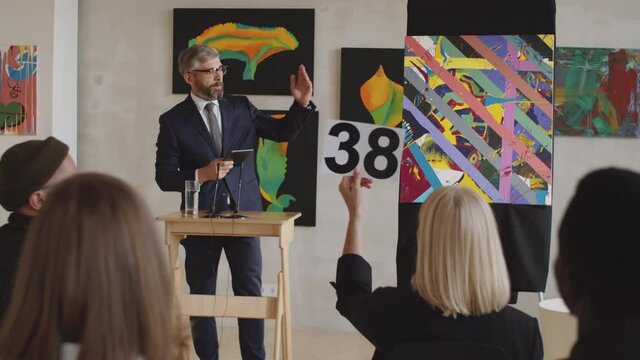 Arc shot of professional male auctioneer in formal suit holding digital tablet, pointing at paddles and calling out bids while selling abstract painting at art auction in gallery