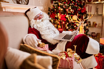 Fototapeta na wymiar Santa Claus sitting at his home and reading email on laptop with сhristmas requesting or wish list near the fireplace and tree with gifts. New year and Merry Christmas , happy holidays concept