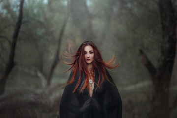 Mysterious fantasy gothic woman dark witch obsessed by evil. Red-haired Girl demon in black dress cape hood. Red hair flutters in wind. Dark dense deep forest background, trees. Scleral lenses on eyes - obrazy, fototapety, plakaty