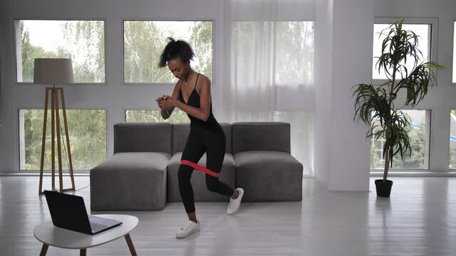 Sporty african american woman online fitness trainer doing cursty lunges with resistance band performing streaming video workout for client amid coronavirus epidemic outbreak, e-learning in quarantine