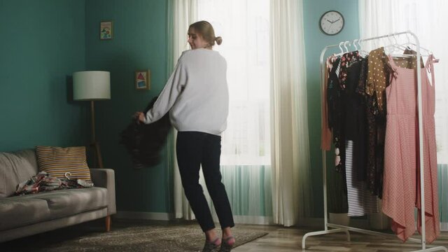 Girl in white sweater and blue jeans stands in middle of room and throws a dress on floor