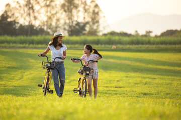 Fototapeta premium Mother and daughter with bicycling at the garden meadow in sunset near white fence. Lifestyle Family Concept