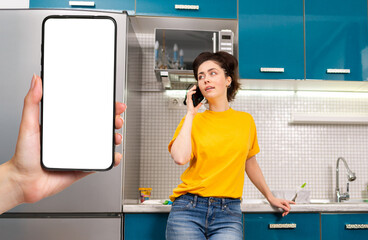 A young beautiful woman communicates on a smartphone in the home kitchen. Female's hand holding a smartphone with mock up. Communication and household concept