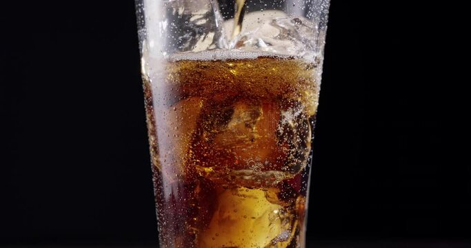 Cola is poured into a glass of cola with ice. The glass stands on a wooden table with a black background. Gas bubbles actively rise in the glass. Slow motion, Blackmagic Ursa Pro G2.