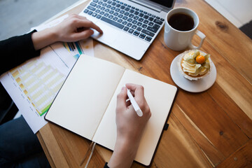 Hands using laptop and notebook with cup of tea and cake on wooden table in cafe