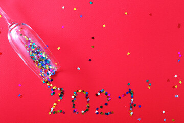 Champagne glass with star shaped sequins scattered in the form of inscription 2021 on a red background.