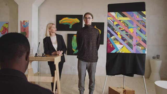 Zoom in of young male artist getting introduced by female auctioneer and presenting his abstract painting to audience at art auction in gallery