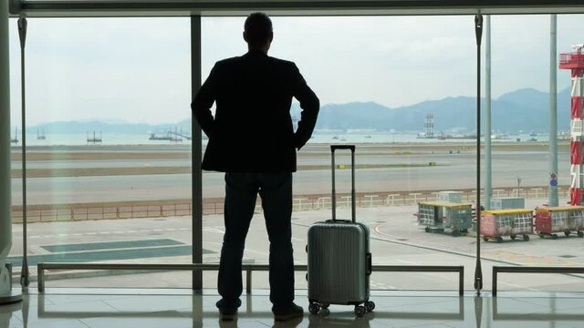 Passenger man stay against concourse window, put hands on hips and look out to empty taxiways of modern airport at reclaimed land, water and mountains on background. Small rollaboard case stand near