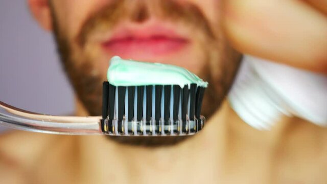 A young bearded man squeezes toothpaste onto a brush and brushes his teeth close-up