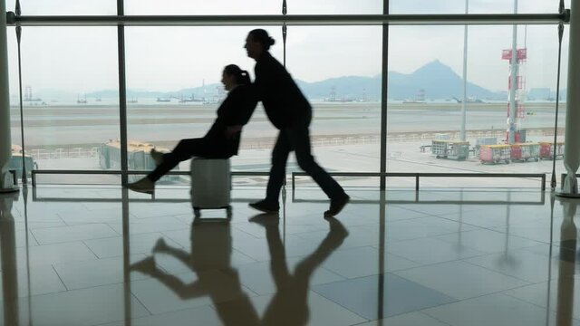 Funny couple at airport, woman race on trolley case, man run behind and push rider forward. Silhouetted shot against terminal window. Passengers have fun after long flight arrival, enjoy like kids