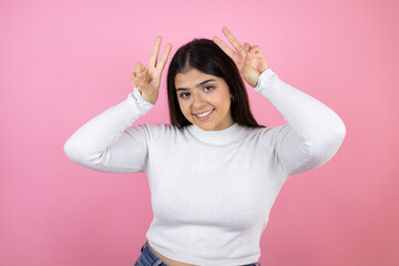 Young beautiful woman over isolated pink background Posing funny and crazy with fingers on head as bunny ears, smiling cheerful