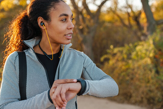 Pleased african american sportswoman in earphones using smartwatch