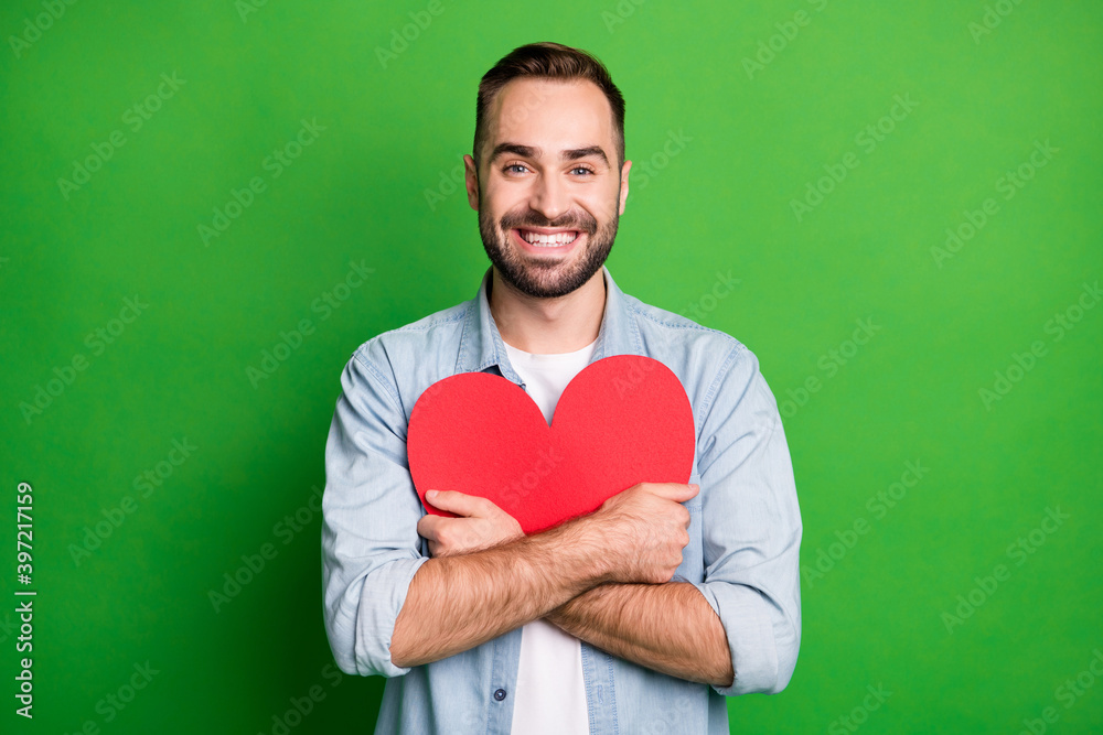 Wall mural Portrait of optimistic guy hug red heart wear blue shirt isolated on vibrant green color background