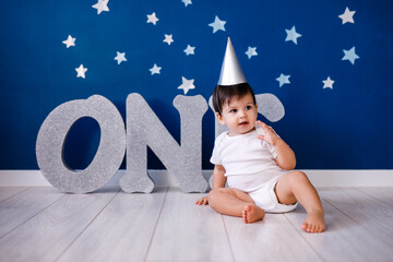 Baby boy of one year old wearing a white body and a festive paper hat sits on the floor on a blue...