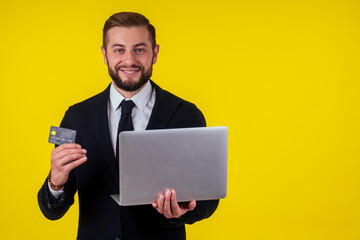 Attractive excited young brunette man isolated over yellow background holding laptop computer and plastic credit card