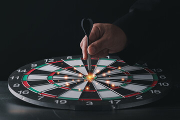 Obraz premium Businessman hand in black suite holding black dart put to center of target board on vintage table meaning of investment and achievement goal. Planning and business strategy concept.