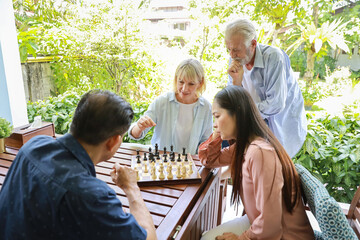 Four multiethnic senior people wearing casual dressed sitting at house, playing chess board and having fun together. Elder caucasian husband man teaching caucasian wife woman how to play.