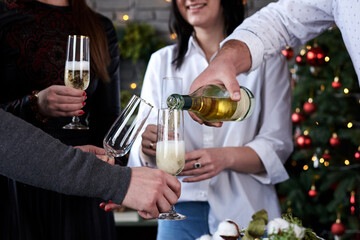 Close-up picture of hands with glasses, and bottle of champagne, Christmas tree on background. New year party celebration with friends and family at home.