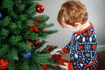 A child with a Christmas tree. A little blond boy  reaches for the toy on a beautiful green Christmas tree. Christmas photo